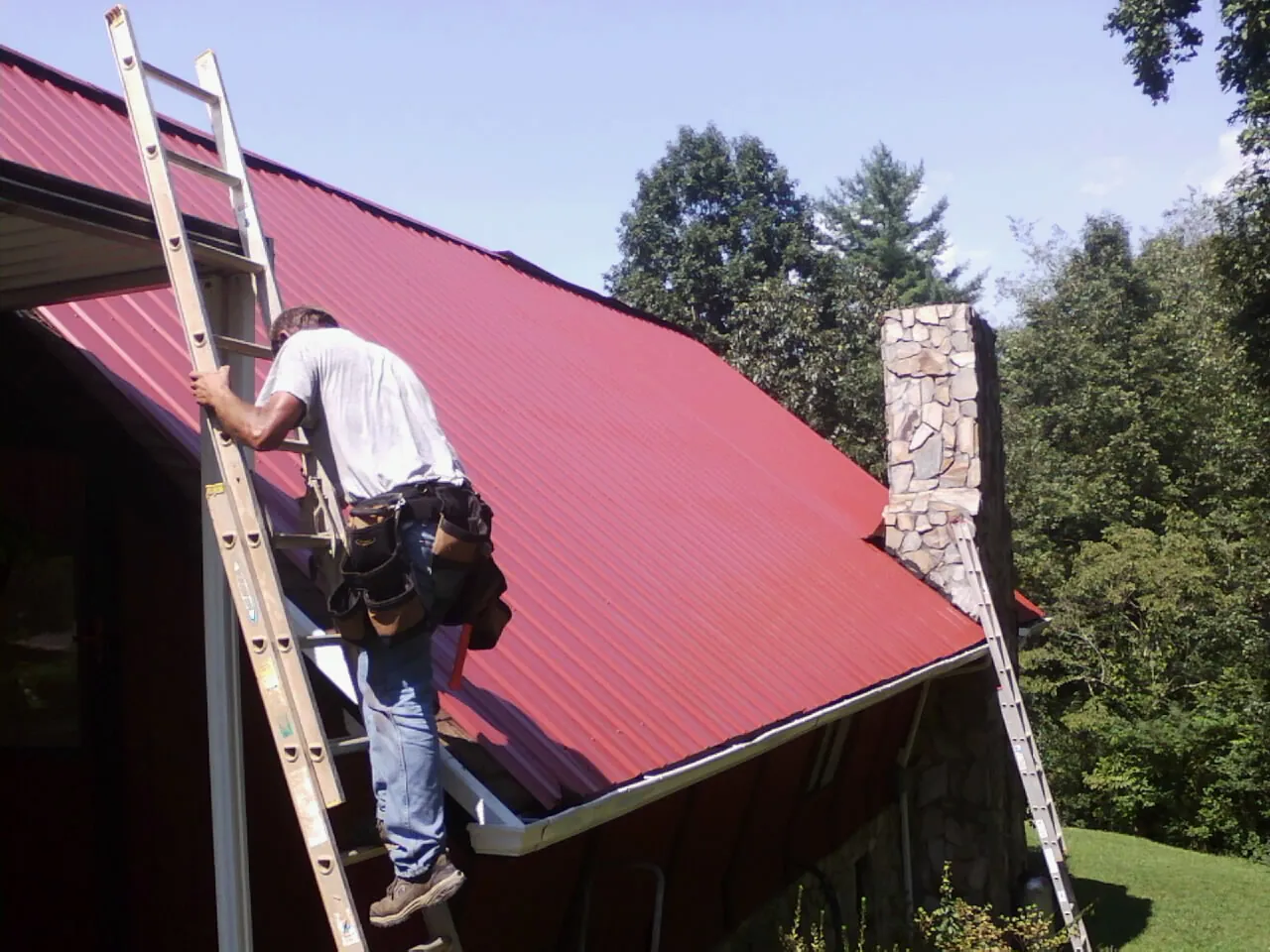 Roofer on a ladder beside a red metal roof and stone chimney — Soffit & Fascia Repair by Roofing Brothers in Belchertown